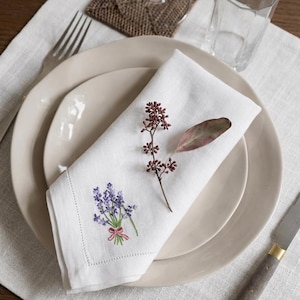 May include: A white linen napkin, embroidered with a lavender bouquet and a pink bow, sits on a beige plate. A sprig of dried berries and a leaf are placed on the napkin. A fork, knife, and glass complete the setting.