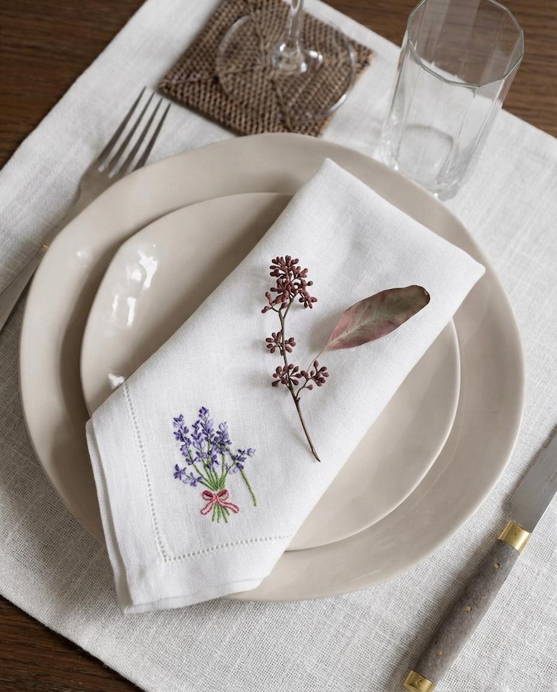 Puede incluir: Una servilleta de lino blanco con un ramo de lavanda bordado y un lazo rosa, colocada sobre un plato beige. Una ramita de bayas secas y una hoja adornan la servilleta. El conjunto incluye un tenedor, un cuchillo y un vaso.