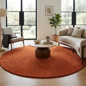 May include: A circular rug in a burnt orange hue, featuring a sunburst pattern, is the focal point of the living room. A light wood coffee table and a beige sofa are placed on the rug. The room also includes a chair, a floor lamp, and a potted plant.