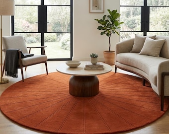 Puede incluir: Una alfombra grande y redonda de color óxido, con un diseño de rayos de sol, domina la sala de estar. La alfombra está centrada debajo de una mesa de centro de madera clara y un sofá beige. La habitación también cuenta con una silla, una lámpara de pie y una planta en maceta.