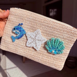 May include: A beige woven clutch with a zipper closure, adorned with beaded embellishments. The embellishments include a blue dolphin, a white starfish with pearl accents, and a turquoise seashell with a pearl. The clutch is held by a person.