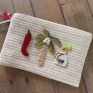 May include: A beige woven clutch bag with a zip closure, decorated with beaded embellishments. The embellishments include a red chilli pepper, a palm tree, and a tequila bottle with the word "TEQUILA".