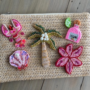 Puede incluir: Bolso de mano de paja tejida adornado con abalorios de temática tropical. Presenta una langosta, una concha, una palmera, una flor y una botella de tequila. Los adornos son rosas, verdes y blancos.
