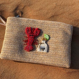 May include: A beige woven clutch bag with a zip, decorated with beaded details. A red lobster and a tequila bottle with a lime slice are attached. The bag is on a sandy surface.