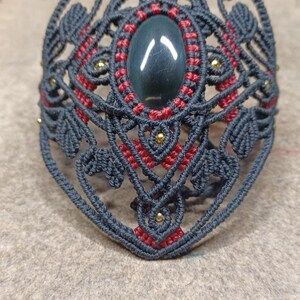 May include: A handmade macramé bracelet in dark grey, with intricate patterns, red detailing, and small gold beads. It features an oval, dark green stone at its centre.