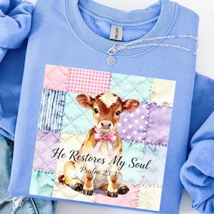 May include: Light blue sweatshirt featuring a graphic of a calf with a pink bow, set against a patchwork quilt design. The text "He Restores My Soul Psalm 23:3" is printed below the calf. A silver chain necklace is draped across the neckline.
