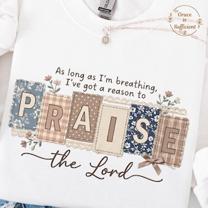 May include: White sweatshirt with the text "As long as I'm breathing, I've got a reason to PRAISE the Lord." The word "PRAISE" is in a patchwork design with floral accents. A necklace with a pendant is visible.