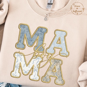 Könnte beinhalten: Beiges Sweatshirt mit dem Wort "MAMA" in großen, verzierten Buchstaben. Die Buchstaben sind goldfarben umrandet und mit einem blauen Blumenmuster gefüllt. Das Wort "boy" steht kursiv zwischen den Buchstaben.