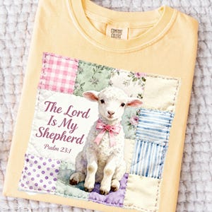May include: Pale yellow Comfort Colors t-shirt with a patchwork design. The patchwork features a lamb with a pink bow and the text "The Lord Is My Shepherd" and Psalm 23:1.