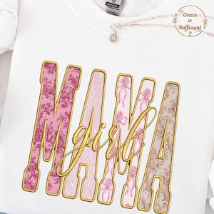 Könnte beinhalten: Weißes Sweatshirt mit dem Wort "MAMA girl" in goldener Schrift. Die Buchstaben sind mit rosa und beigen floralen Mustern gefüllt. Eine Halskette mit Anhänger ist sichtbar.