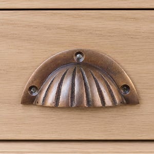 Puede incluir: Un tirador de cajón en forma de media luna con acabado en bronce oscuro y una textura acanalada similar a una concha. El herraje presenta tres orificios visibles para tornillos de montaje y está fijado sobre una superficie de madera clara con veta horizontal.
