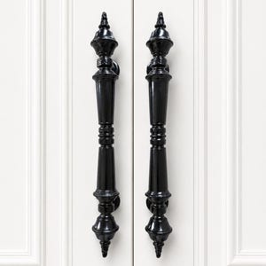 May include: Two vertical black door handles with a glossy finish and a decorative, multi-tiered spindle design. The handles are mounted on a pair of white cabinet doors featuring recessed paneling and vertical molding details.