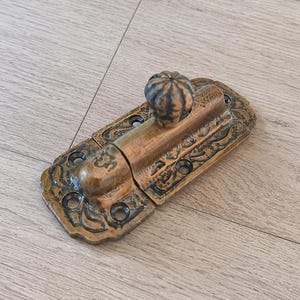 May include: A rectangular brass window sash lock featuring an antique finish with dark patina accents. The hardware includes a central fluted knob and a decorative embossed scroll pattern along the base plate, which contains six pre-drilled screw holes.