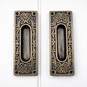 Puede incluir: Dos tiradores empotrados de color bronce antiguo. Cada tirador rectangular presenta un agarre ovalado para los dedos y diseños florales y de volutas simétricos y ornamentados. Los tiradores están montados sobre una superficie blanca.
