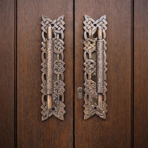 Könnte beinhalten: Bronzefarbene Türgriffe mit aufwendigen keltischen Knotenmustern. Die Griffe sind an einer dunkelbraunen Holztür befestigt, mit einem Schlüsselloch. Die Griffe haben einen strukturierten Griff und dekorative Platten.