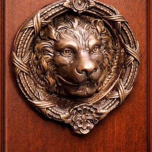 Puede incluir: Un aldaba de puerta de bronce con forma de cabeza de león con detalles intrincados, colocada sobre una puerta de madera oscura. La aldaba presenta una cara de león detallada dentro de un marco decorativo, agregando un toque de elegancia clásica.