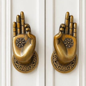 May include: Two antique brass door handles shaped like hands in a gesture of blessing. Each hand features a detailed floral design on the palm and is mounted on a circular base with an ornate pattern. The handles are set against a white door.