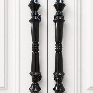 May include: Two black, ornate door pulls with a glossy finish. Each pull features a decorative finial at the top and bottom, with a series of rounded and tapered sections. The pulls are mounted on a white door.
