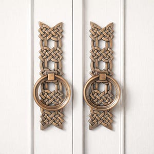 May include: Two bronze-colored cabinet pulls with a Celtic knot design. Each pull features a rectangular backplate with an intricate woven pattern and a circular ring handle. The pulls are mounted on a white cabinet door.