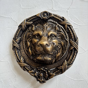 May include: An antique-style bronze lion head door knocker. The lion's face is intricately detailed with a textured mane and facial features. The knocker is framed by a woven border and a floral accent. The background is a textured white surface.