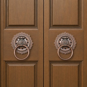 May include: Two matching copper-toned metal door knockers with circular rings and ornate, scalloped backplates mounted on a pair of brown wooden cabinet doors featuring recessed rectangular panels and a vertical seam down the center.