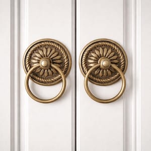May include: Two antique brass cabinet pulls. Each pull features a circular backplate with a floral design and a rope-like border. A round ring handle hangs from the center. The pulls are mounted on a white cabinet door.