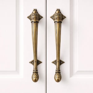 Könnte beinhalten: Zwei antike Messing-Schrankgriffe mit aufwendigen Details. Jeder Griff hat eine dekorative Oberseite, einen langen, geriffelten Griff und eine ananasförmige Basis. Die Griffe sind an einer weißen Schranktür montiert.