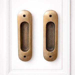 Może przedstawiać: Dwa antyczne, wpuszczane uchwyty do drzwi z mosiądzu. Każdy owalny uchwyt ma prostokątny, wpuszczony obszar chwytu. Zamontowane na białych drzwiach. Mosiądz ma ciepłą, postarzaną patynę.