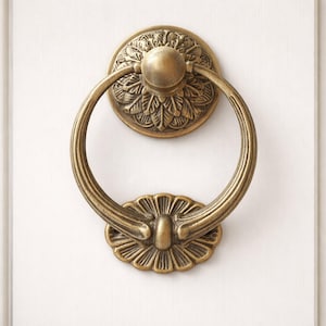 May include: Ornate brass door knocker with a circular handle and decorative floral accents. The knocker is mounted on a white door, showcasing its antique design. The door knocker's ring is approximately 4 inches in diameter.