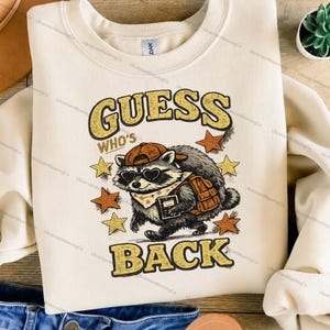 May include: A cream-colored crewneck sweatshirt featuring a graphic of a raccoon wearing a baseball cap, heart-shaped sunglasses, and a backpack. The raccoon holds a book. The text reads GUESS WHO'S BACK in a distressed yellow font with stars.