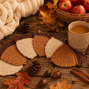 May include: A set of five crocheted acorn-shaped coasters arranged in a fan pattern, featuring alternating shades of cream, tan, and brown yarn with dark brown caps. The coasters are textured with a visible stitch pattern and have a rounded shape.