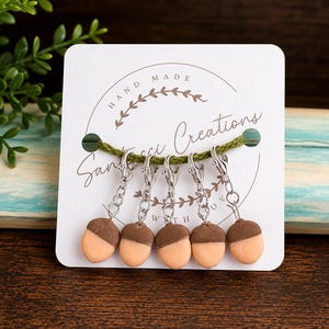 May include: Five acorn-shaped stitch markers attached to a white display card with the text "Hand Made" and "Sanucci Creations". Each marker features a brown cap and a light peach-colored base, connected by a silver-toned metal lobster clasp.