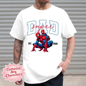 May include: White t-shirt featuring a graphic of two Spider-Man characters, one adult and one child, in red and blue. The word "DAD" is above the image in blue block letters, with "super" in red script. The text "EST. 2026" is also present.