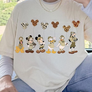 Puede incluir: Camiseta beige con personajes de dibujos animados: Pluto, Mickey Mouse, Minnie Mouse, Pato Donald y Goofy con trajes de safari. Cada personaje sostiene un globo con forma de cabeza de Mickey Mouse y estampados de animales. La camiseta tiene una camiseta interior blanca de manga larga.