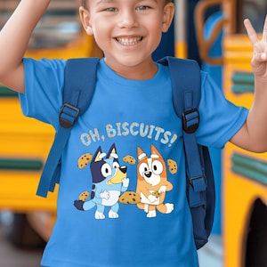 Könnte beinhalten: Ein hellblaues T-Shirt mit der Aufschrift "OH, BISCUITS!" und Cartoon-Hundfiguren. Das T-Shirt hat einen Rundhalsausschnitt und kurze Ärmel. Die Cartoon-Figuren sind ein blauer und ein orangefarbener Hund, umgeben von Keksen.