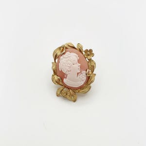 Puede incluir: Broche vintage con camafeo que presenta el perfil de una mujer en blanco sobre un óvalo melocotón. El marco de metal dorado tiene detalles de hojas y flores. Un accesorio clásico y atemporal para un toque de elegancia.