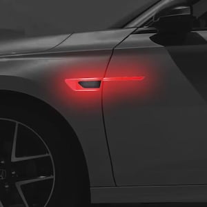 Pode incluir: Um painel lateral de carro prateado com uma faixa de luz horizontal vermelha brilhante integrada na ventilação do para-lama dianteiro. O veículo possui um retrovisor escuro e parte de uma roda de liga leve visível na base.