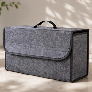 May include: A rectangular dark gray felt storage box with a structured design, featuring a black fabric handle on the top lid and black trim along the edges. The container has a flap closure that covers the front, providing a clean and organized look.