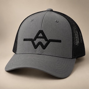 May include: A charcoal grey trucker hat featuring a black mesh back and a solid front panel. The front displays a black embroidered logo consisting of the letters A and W connected by a horizontal line, with a curved brim and stitched detailing.