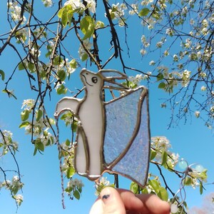 May include: A stained glass pterodactyl suncatcher with a white body and iridescent blue wings. The suncatcher is held in front of a blue sky and flowering tree branches.