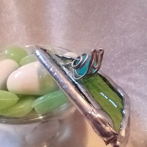 May include: A handcrafted, stained glass piece with a silver-colored metal frame. The glass is a vibrant green with a turquoise accent. The piece is next to a glass jar filled with white and green candies.