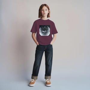 Puede incluir: Una camiseta granate con un gráfico de una luna y el texto "THE NEXT LEAP" y "SENTIENCE". La camiseta se lleva con vaqueros azul oscuro y zapatillas de deporte color canela.
