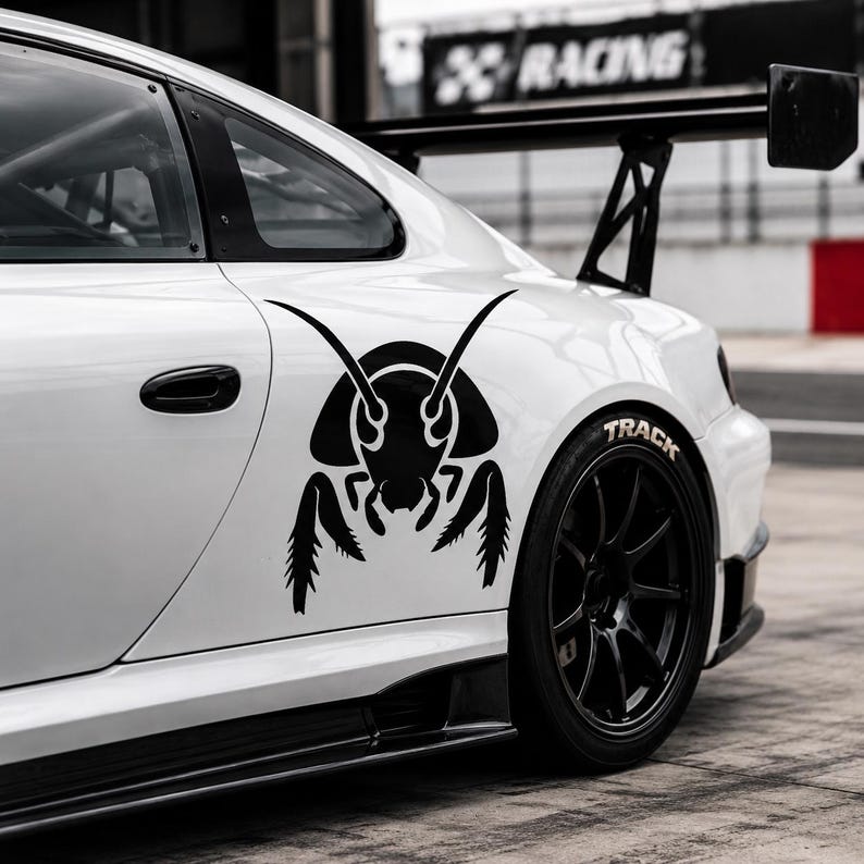 Op de afbeelding: Een witte sportwagen met een grote zwarte insectensticker op het zijpaneel. Het voertuig is voorzien van een zwarte achterspoiler, zwarte meerspaaks wielen met banden waarop TRACK staat en een zwarte deurgreep op een verhard oppervlak.