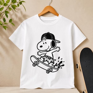 Puede incluir: Camiseta blanca con un gr&aacute;fico negro de Snoopy montando en monopat&iacute;n. Snoopy lleva una gorra de b&eacute;isbol al rev&eacute;s y un ba&ntilde;ador. El monopat&iacute;n tiene ruedas negras. La camiseta est&aacute; colgada en una percha de madera.