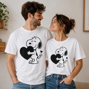 Puede incluir: Dos camisetas blancas con un perro de dibujos animados en blanco y negro sosteniendo un coraz&oacute;n negro. El perro tiene una cara sonriente y un peque&ntilde;o coraz&oacute;n al lado. Las camisetas son usadas por dos personas.