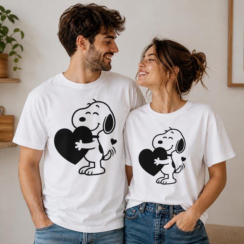 Puede incluir: Dos camisetas blancas con un perro de dibujos animados en blanco y negro sosteniendo un coraz&oacute;n negro. El perro tiene una cara sonriente y un peque&ntilde;o coraz&oacute;n al lado. Las camisetas son usadas por dos personas.
