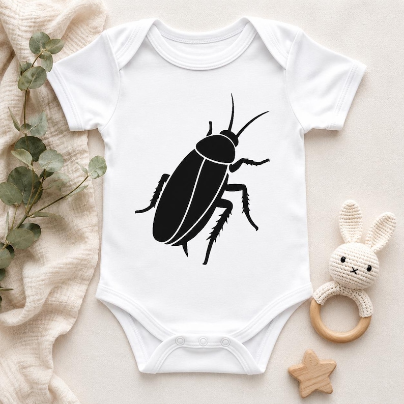 Op de afbeelding: Een wit babyrompertje met korte mouwen en een grote, zwarte afbeelding van een kakkerlak in het midden op de voorzijde. Het kledingstuk heeft een ronde halslijn en drie drukknopen aan de onderkant voor gemakkelijke toegang, gemaakt van witte stof.