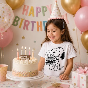 Puede incluir: Una persona joven con una camiseta gr&aacute;fica de Snoopy y un gorro de fiesta sonr&iacute;e a una tarta de cumplea&ntilde;os con velas encendidas. La tarta est&aacute; sobre un soporte blanco para tartas, y el fondo incluye globos y una pancarta de "Feliz Cumplea&ntilde;os".