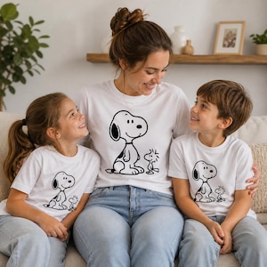 Puede incluir: Tres personas con camisetas blancas con un dise&ntilde;o de dibujos animados de Snoopy y Woodstock en blanco y negro. El gr&aacute;fico de Snoopy est&aacute; centrado en la parte delantera de cada camiseta. Est&aacute;n sentados en un sof&aacute; de color claro.
