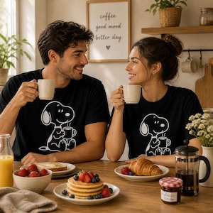 Puede incluir: Dos personas con camisetas negras con un gr&aacute;fico blanco de Snoopy, sosteniendo tazas y sonri&eacute;ndose. Una mesa est&aacute; puesta con art&iacute;culos de desayuno, incluyendo panqueques, fruta y tostadas. Un letrero en la pared dice "coffee good mood better day."